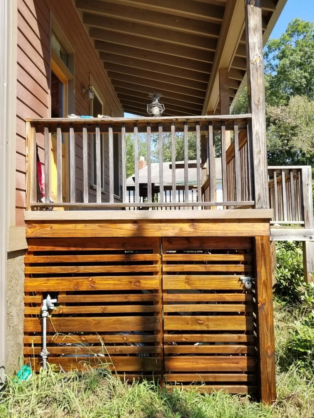 Backyard wooden deck with railing and a gate, attached to a house with brown siding and small windows, surrounded by greenery and mature trees.