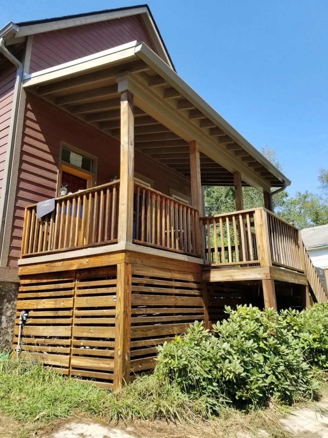 A two-story house with a wooden deck and railing, with stairs leading up to the deck. The house has red siding, a sloped roof, and a clear blue sky overhead.