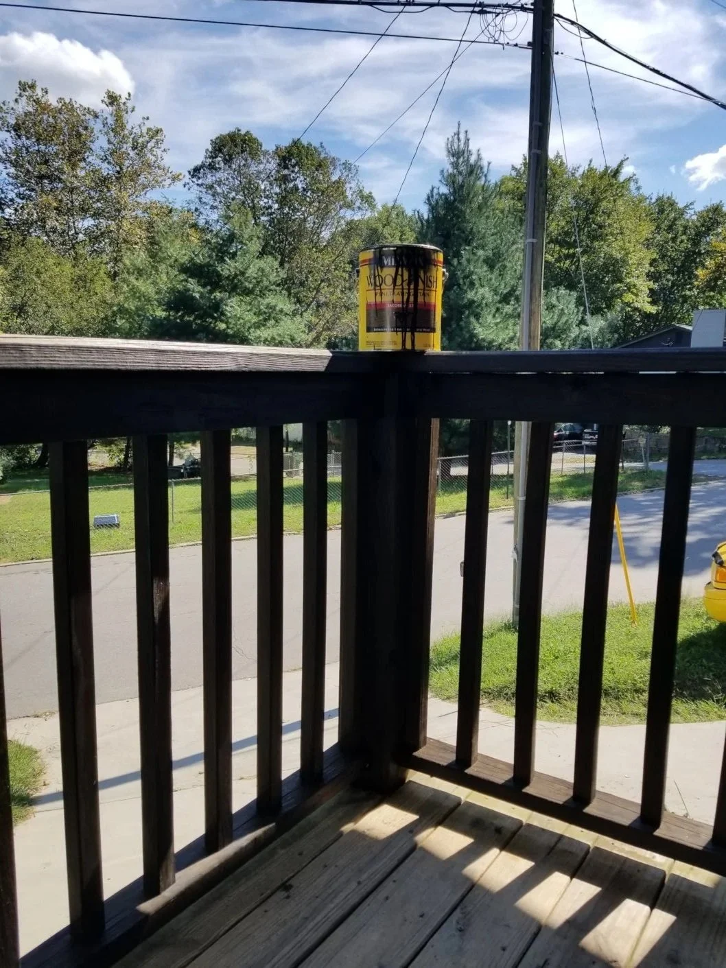View from a wooden balcony with a yellow can on the railing, overlooking a street with trees, a utility pole, a yellow car, and a fence under a blue sky with some clouds.