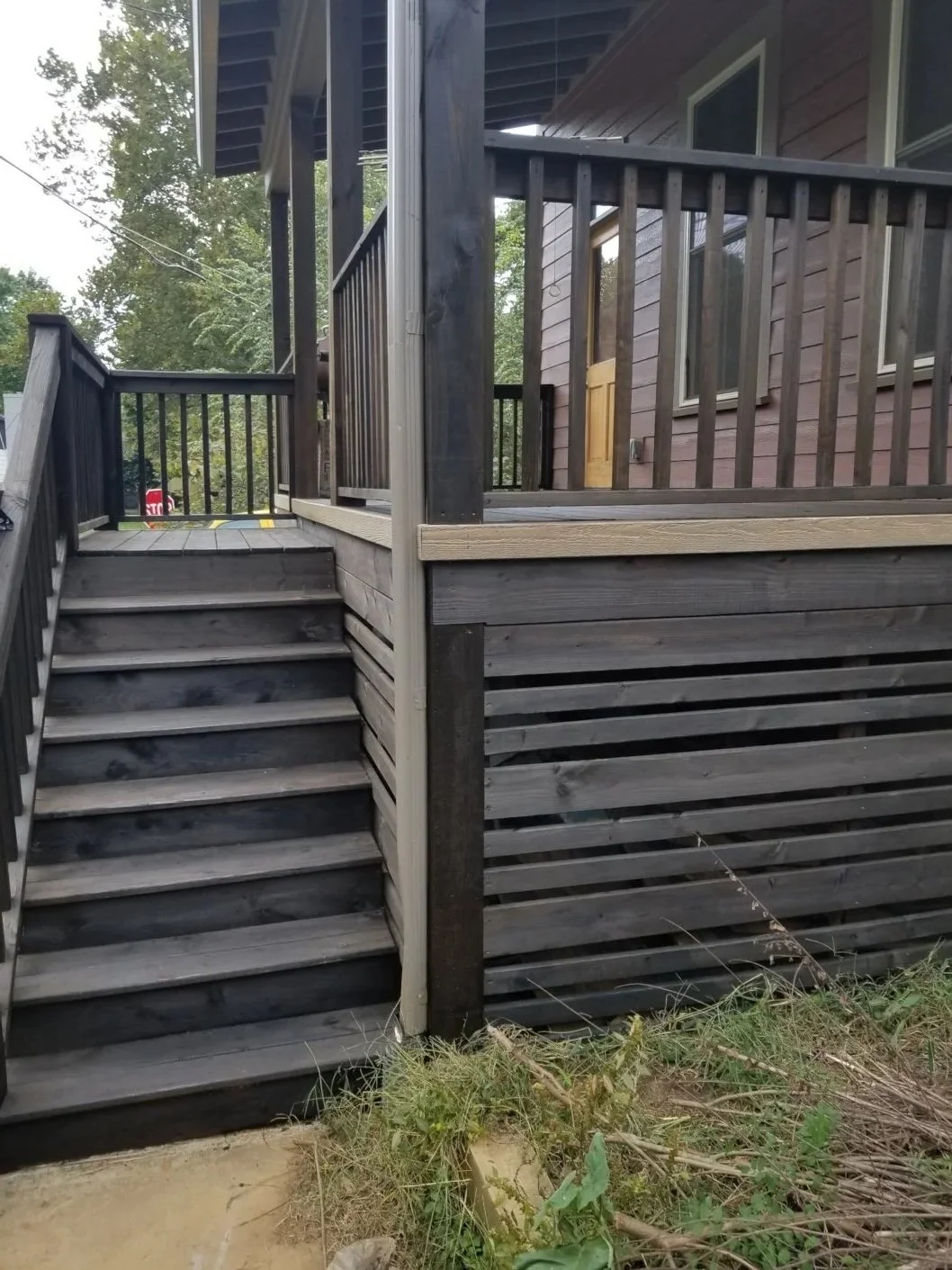 Wooden staircase leading up to a house with a porch and railing, with some grass and plants at the bottom.
