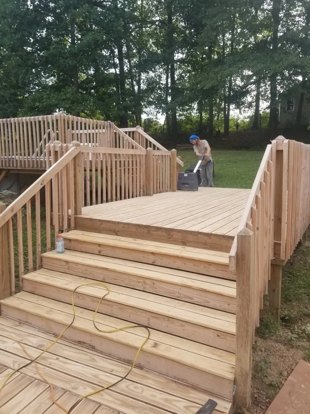 A person in a blue helmet using a power sander on a wooden deck in a backyard with trees in the background.
