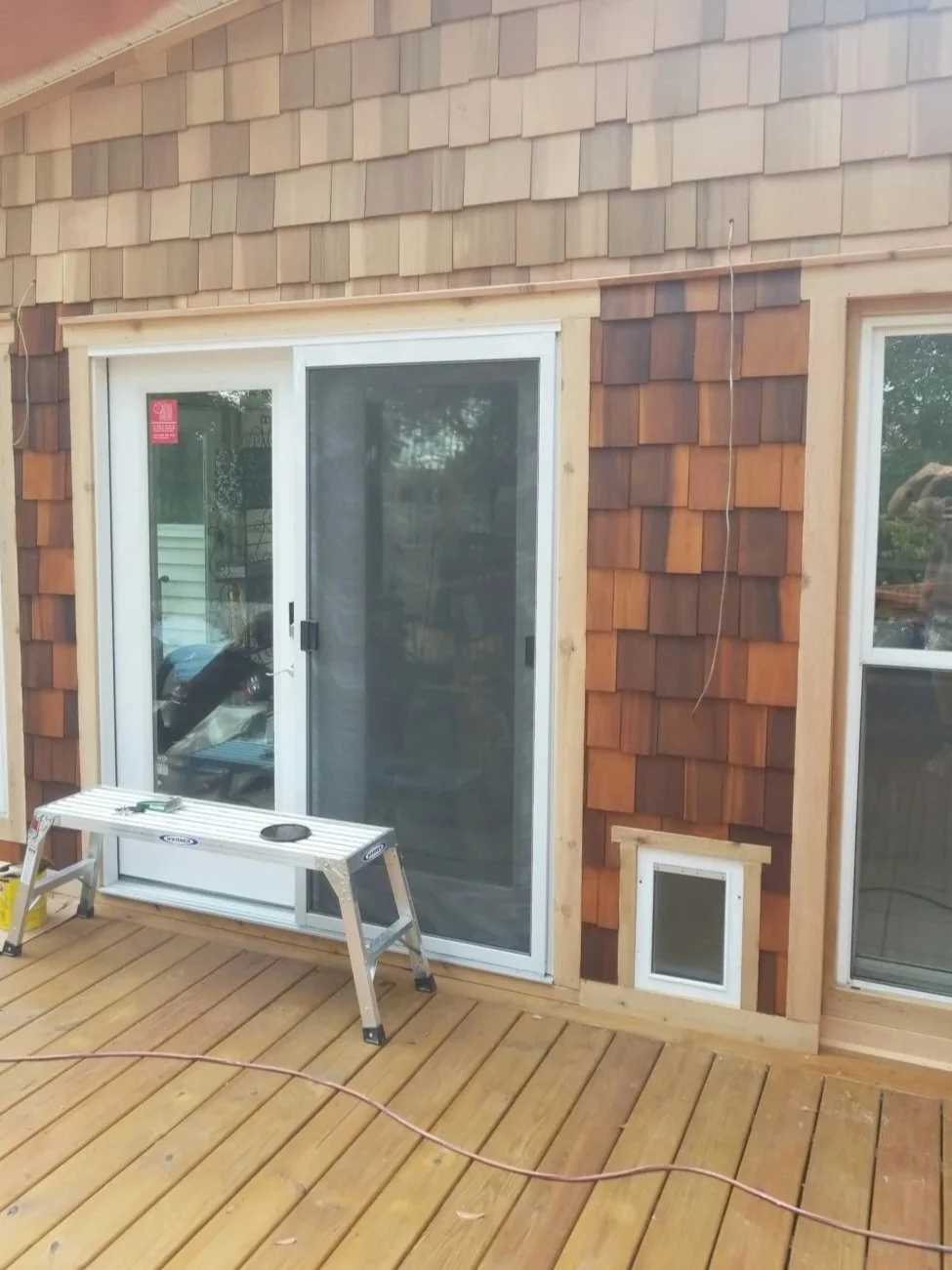 A sliding glass door with a screen, on a wooden deck, in front of a house with wood shingle siding. There is a small white vent in the wall and a step ladder on the deck.