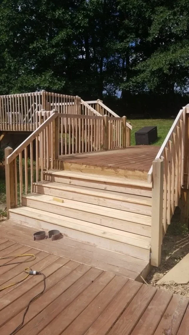 Newly constructed wooden deck with stairs and railings, with some tools and materials on the deck, edges of a grassy yard, and trees in the background.