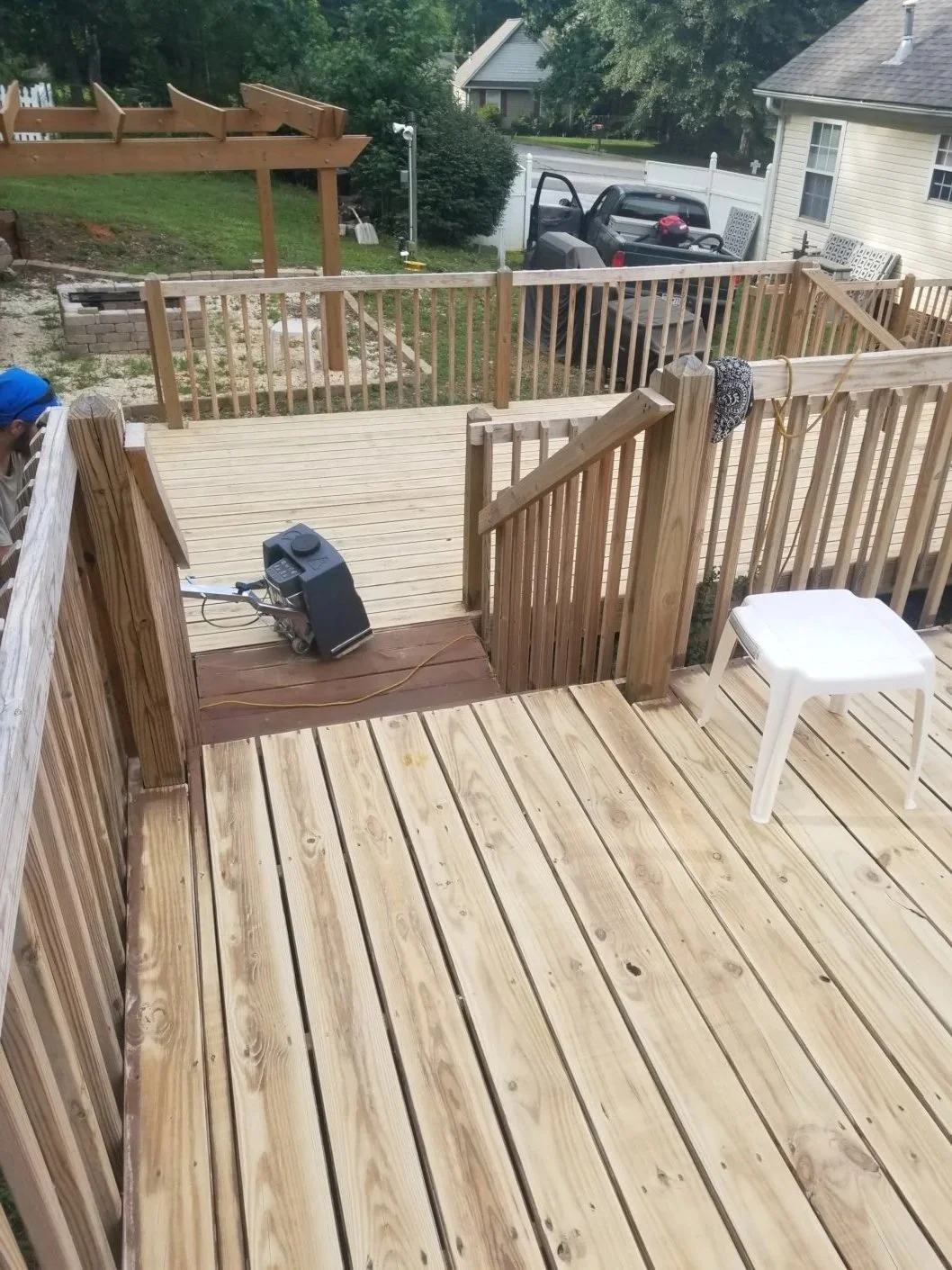 View of a newly built wooden deck with a railing, a white plastic stool, and a heating or cooling device on the deck. The background shows another deck area, a house, a pickup truck parked nearby, and a backyard with grass and trees.