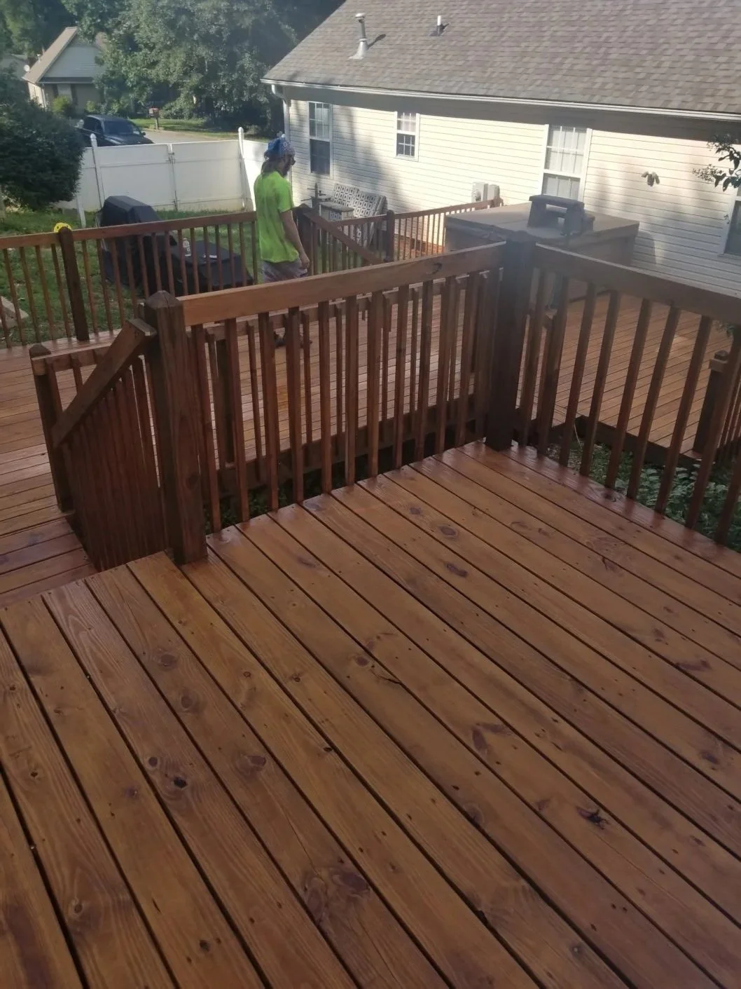 View of a newly stained wooden deck with a railing, in a suburban backyard. A person wearing a bright green shirt and a bandana is standing on the deck near a grill and an outdoor sink.