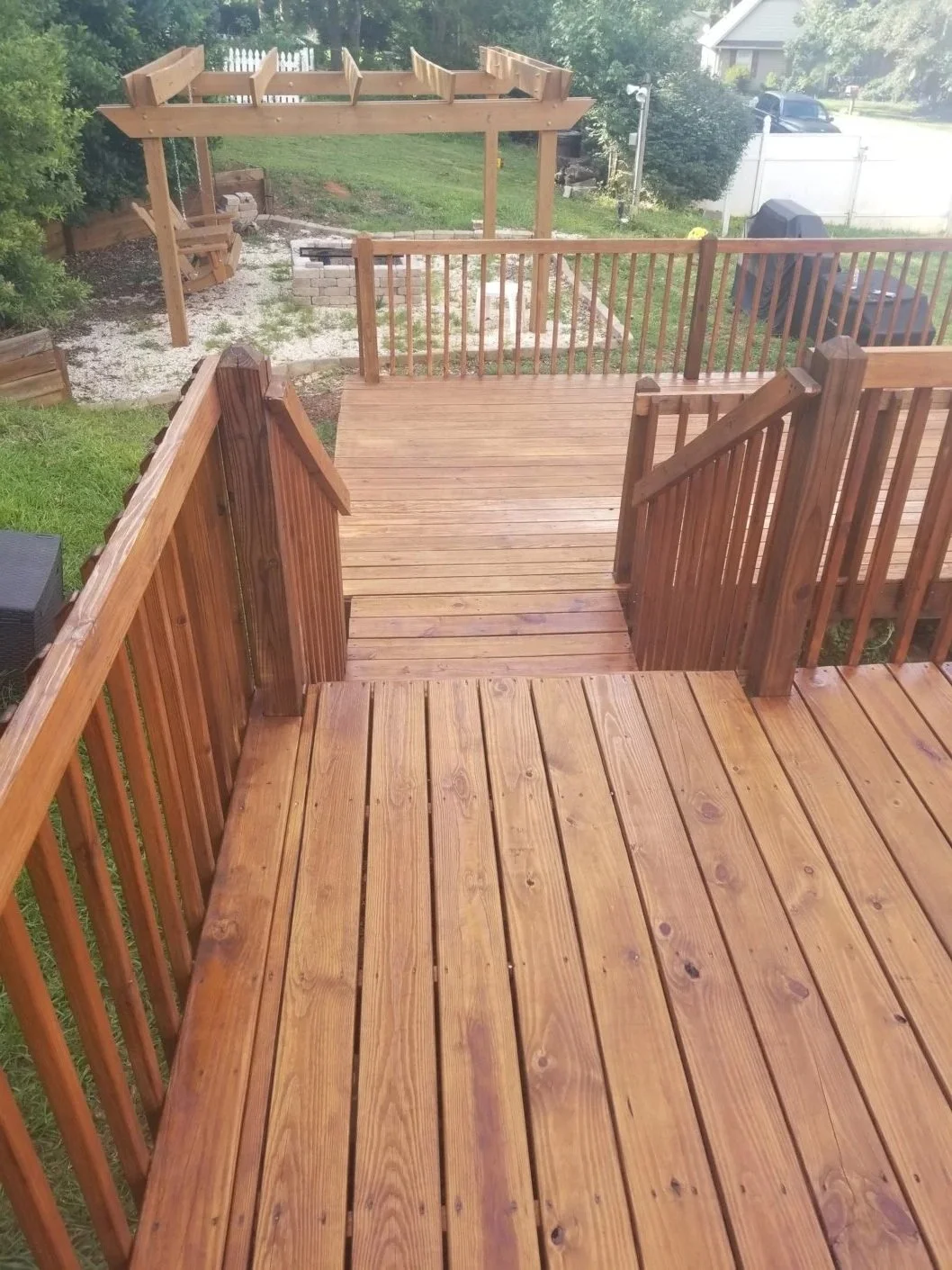 View of a backyard wooden deck with railing, stairs, and a pergola in the background. There is a grassy lawn and a patio area with outdoor furniture.