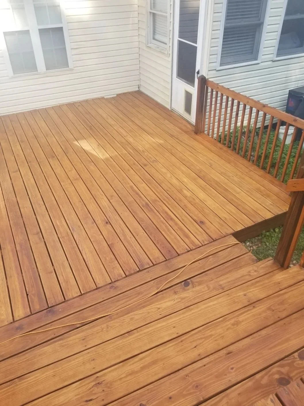 Newly installed wooden deck attached to a white house with multiple windows and a door. Railing around the deck and some electrical cord on the floor.