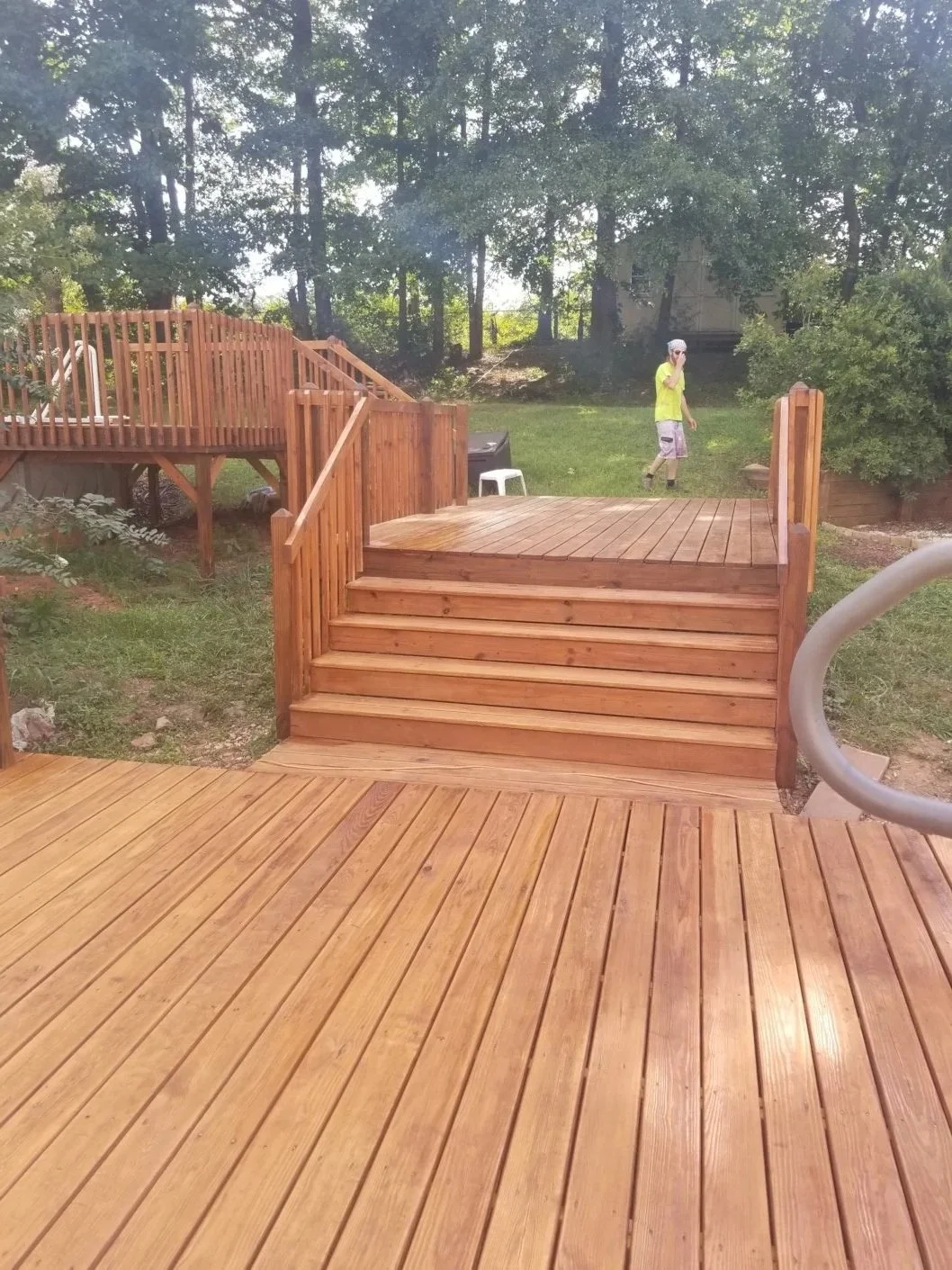 Newly built wooden deck with stairs and railings in a backyard surrounded by trees, with a person walking in the background.