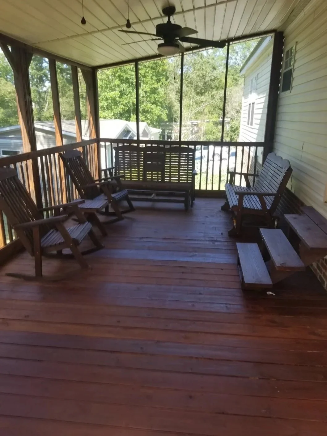  screened-in porch with wooden flooring, ceiling fan, ceiling lights, wooden patio furniture including two chairs, a loveseat, and a small table. Green trees and neighboring houses are visible outside.