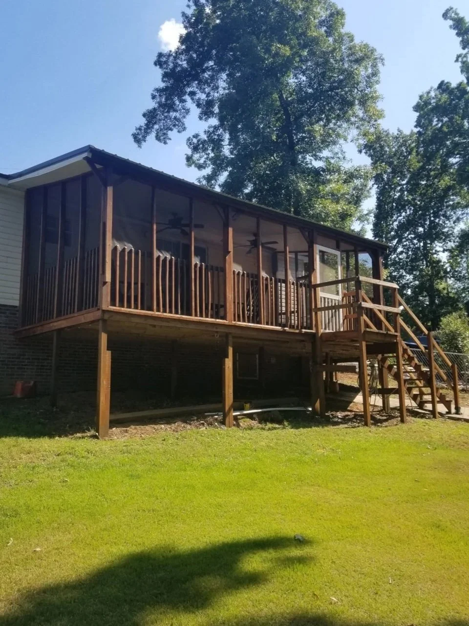 A house with a screened-in porch on stilts, with stairs leading up to it, surrounded by a green lawn and trees.