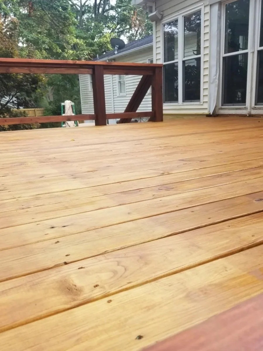 Wooden deck attached to a house with sliding glass doors and a white exterior. Trees and outdoor items are visible in the background.