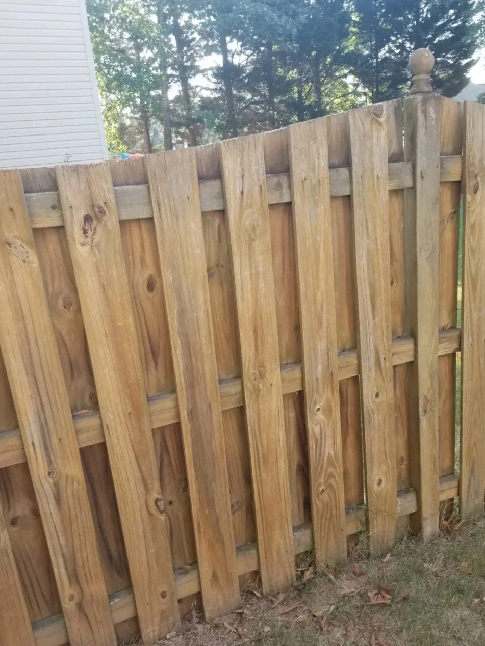 A close-up of a wooden fence with vertical planks and a decorative post on the right side, with trees and a house partially visible in the background.