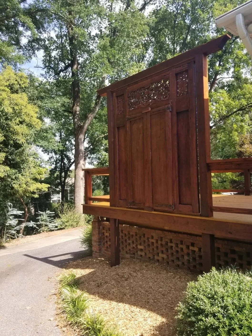 Wooden privacy screen or fence panel on a deck area surrounded by green trees and shrubs under a sunny sky.