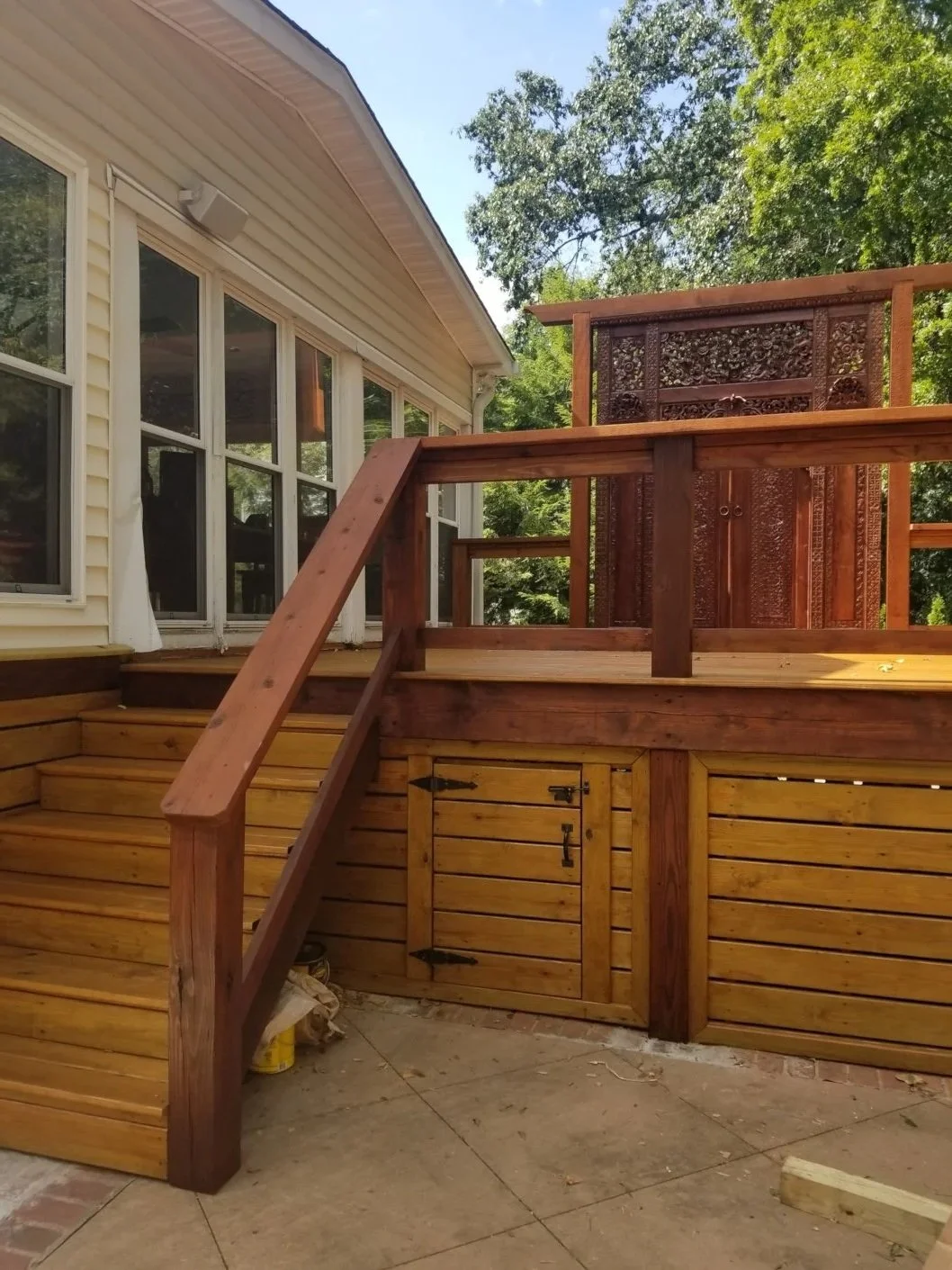 Wooden deck stairs and railing attached to a house with large windows, with trees and blue sky in the background.