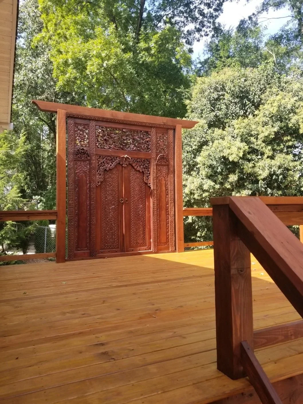 A wooden patio or deck with a decorative wooden door or panel, surrounded by greenery and trees.