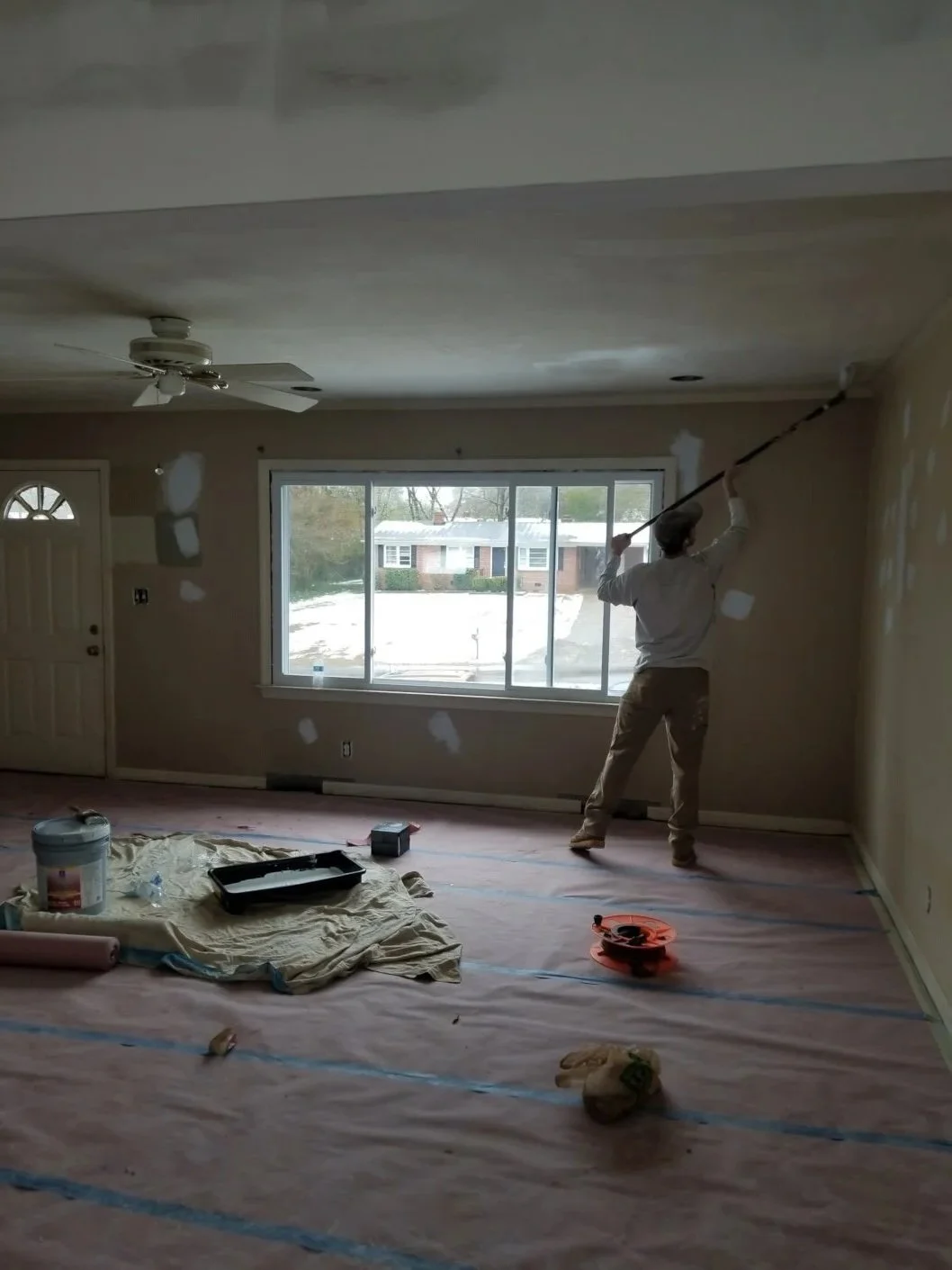 A person sanding a wall in a room under renovation, with supplies and tools on the floor, and a large window showing a snowy outdoor scene.