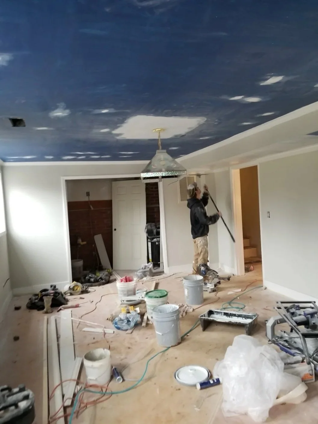 A person painting a ceiling dark blue in a room under renovation, surrounded by construction tools and materials.