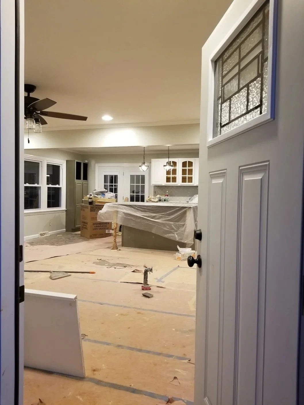 Interior view of a house under renovation, showing a kitchen with construction materials and tools scattered on the floor, covered cabinetry, and unfinished flooring.