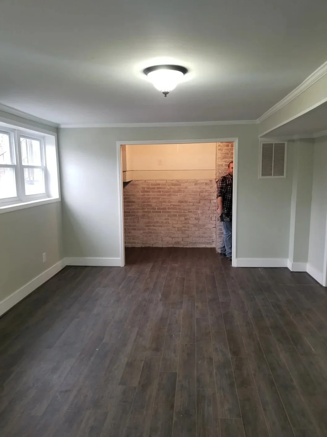 Empty living room with dark wood flooring, light-colored walls, and white crown molding. There are four small windows near the ceiling on the left. A doorway with white trim leads to a brick-walled room where a person is partially visible.