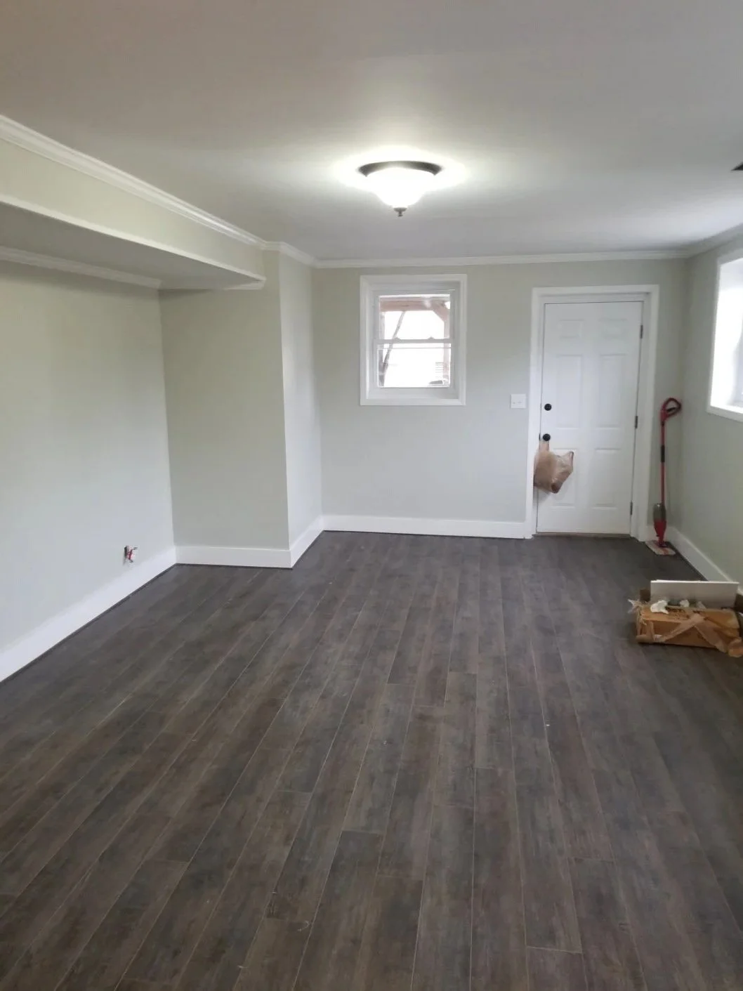 Empty living room or small space with hardwood flooring, light gray walls, white trim, small window, white door, and ceiling light fixture; some construction or renovation materials on the floor.