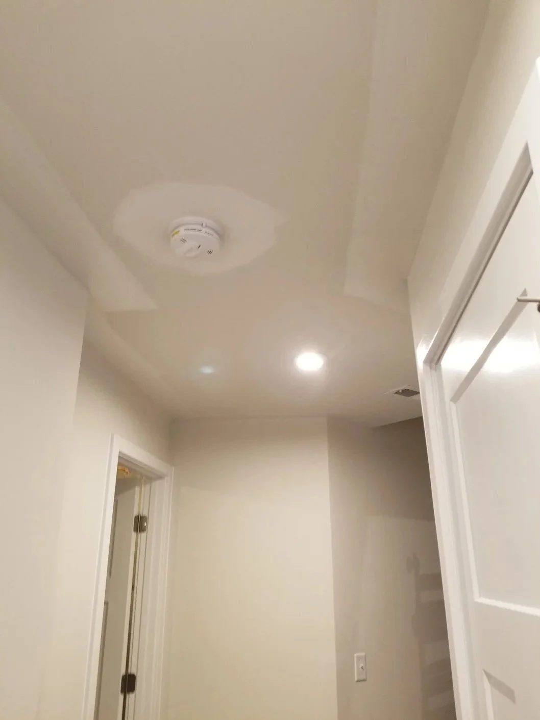 Ceiling with smoke detector, recessed lighting, and air vent, in a hallway with white walls and doorframes.