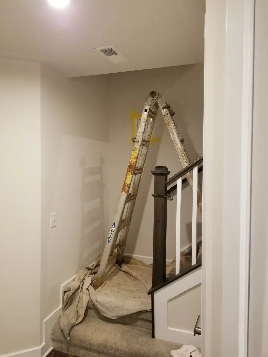 A ladder leaning against a wall in an indoor space, with painter's drop cloths on the floor, indicating ongoing painting or renovation work.
