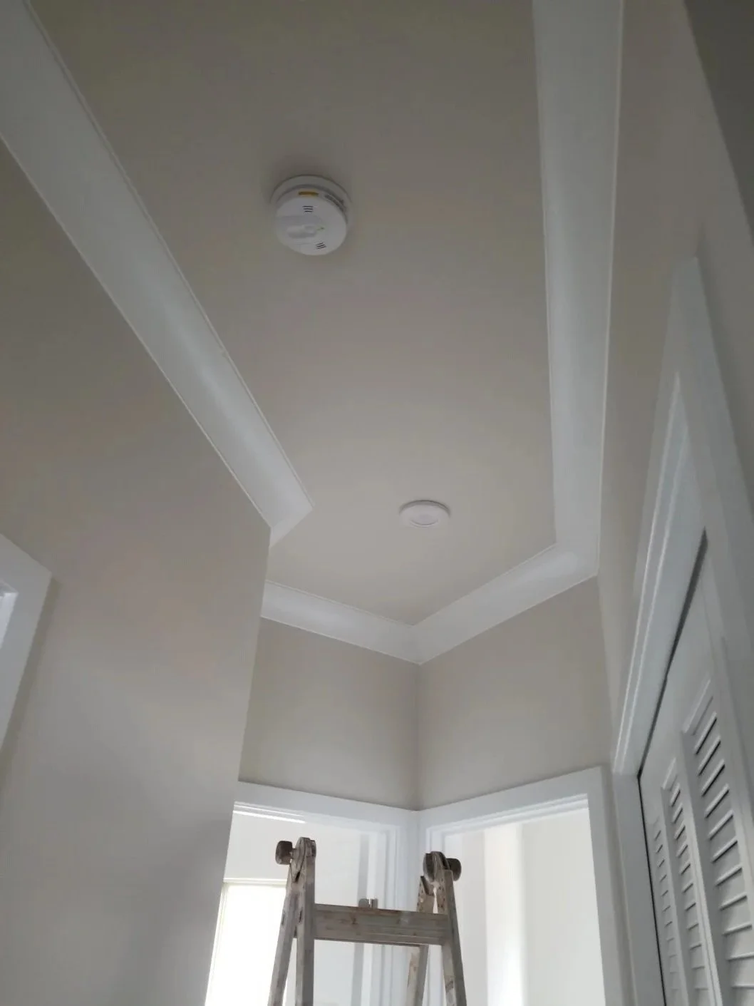 View of a ceiling with a smoke detector, a vent, and recessed lighting, with a ladder and a corner of a room visible.