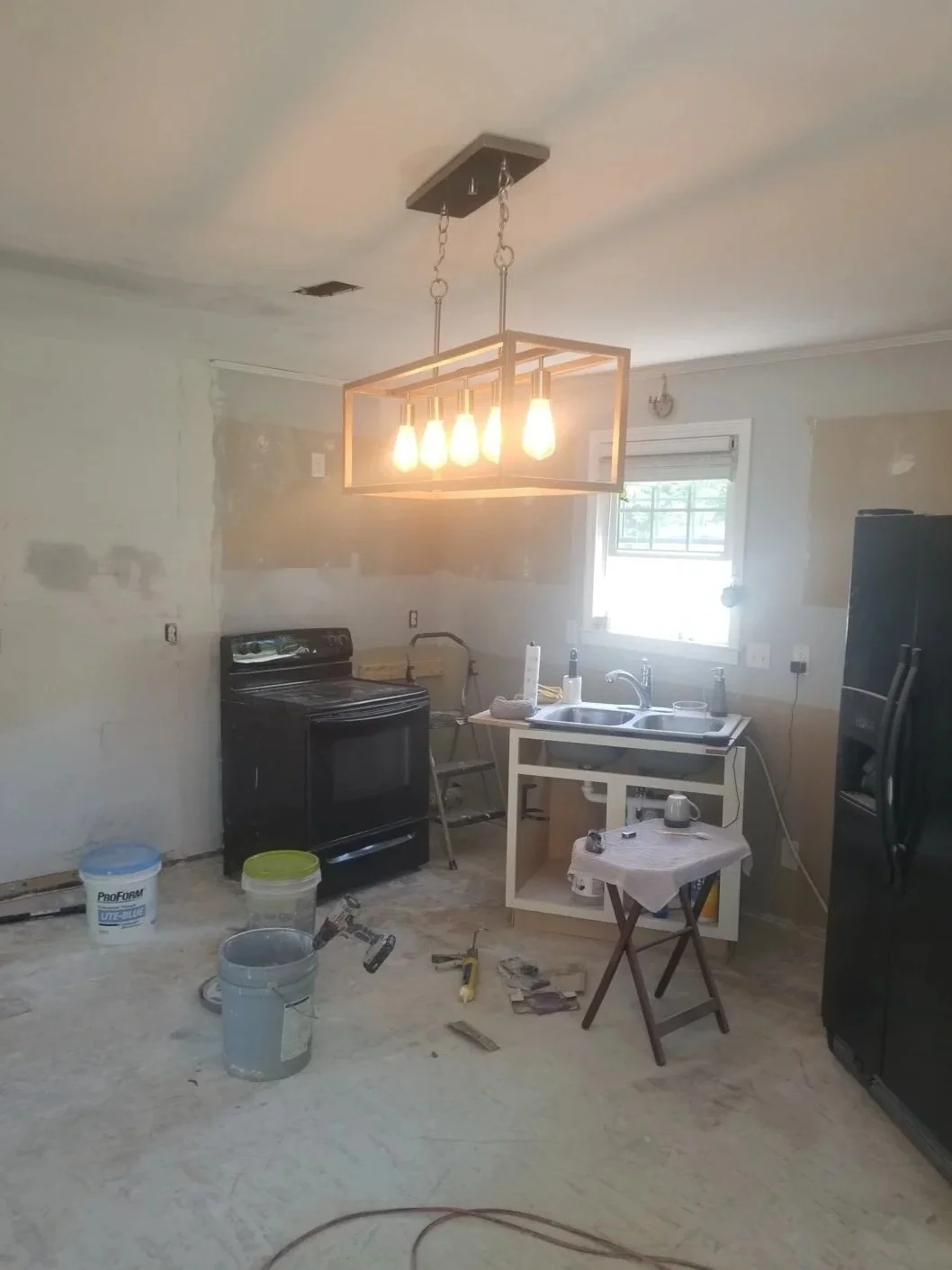 Kitchen under renovation with unfinished walls, a black stove, a white sink, a black refrigerator, construction tools, and a modern rectangular chandelier with exposed bulbs hanging from the ceiling.