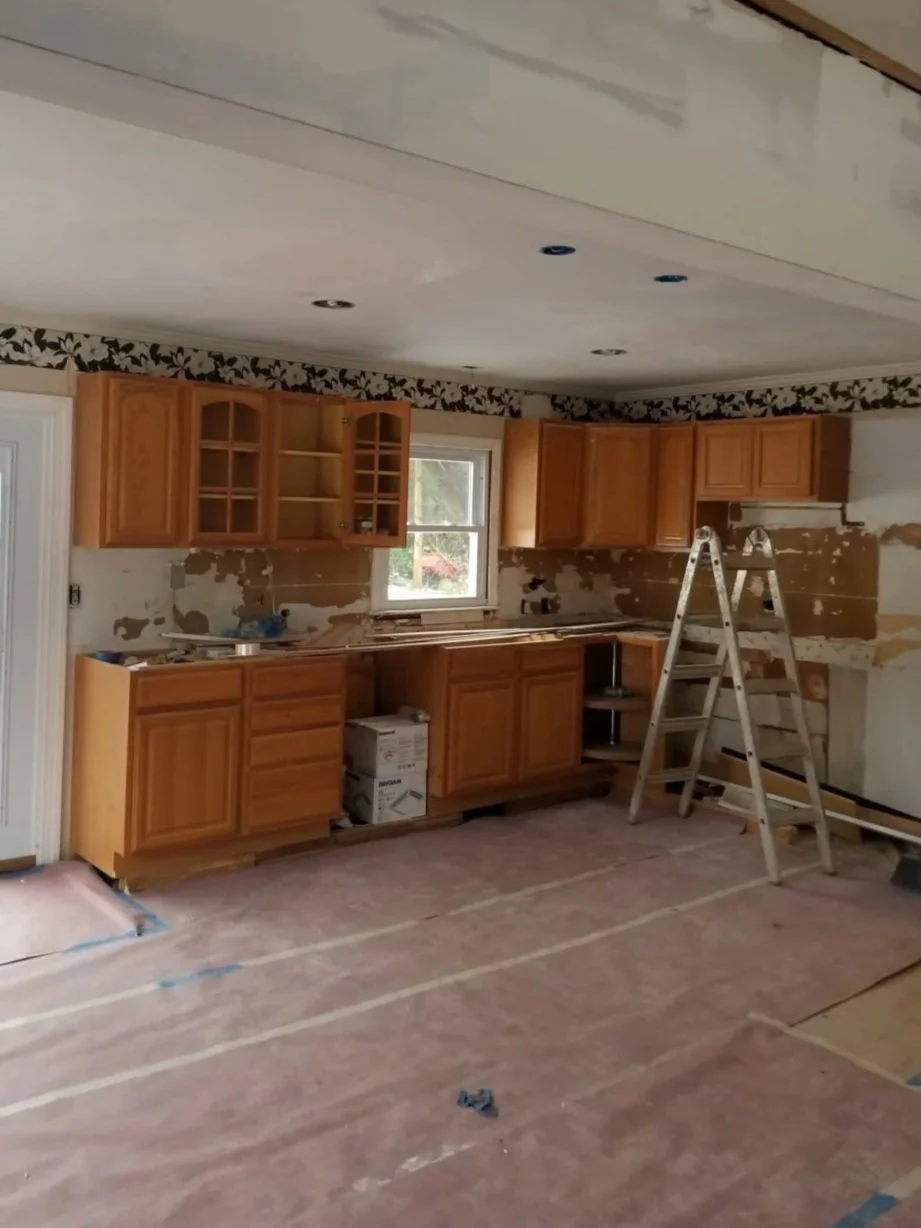 Kitchen undergoing renovation with unfinished walls, upper and lower wooden cabinets, a window, a step ladder, and a cardboard-covered floor.