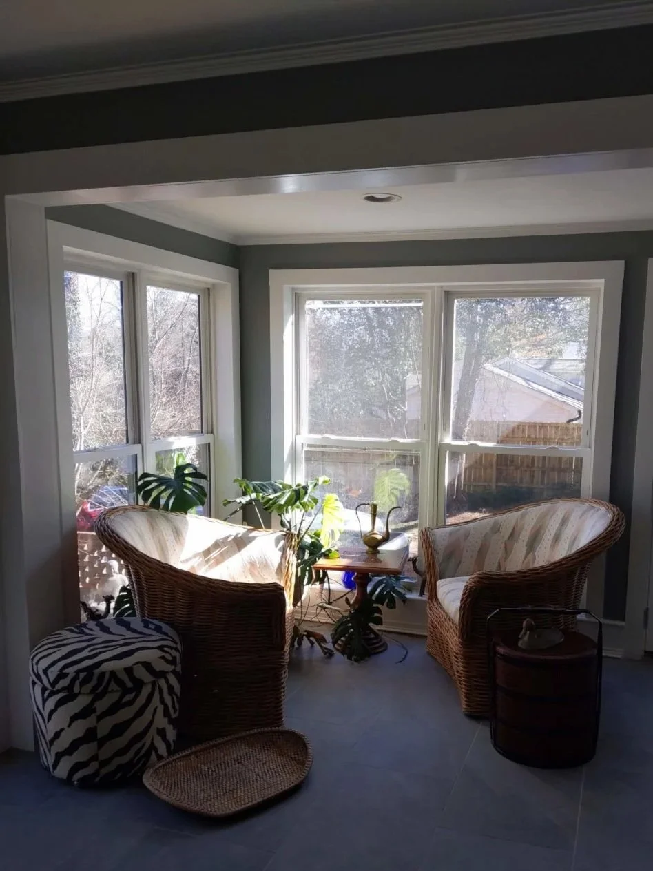 Sunlit enclosed porch with wicker chairs, potted plants, and small side table, overlooking trees and backyard.