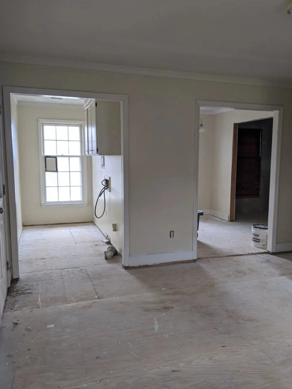 Empty room under renovation with unfinished flooring, a window on the back wall, and construction materials and tools visible.