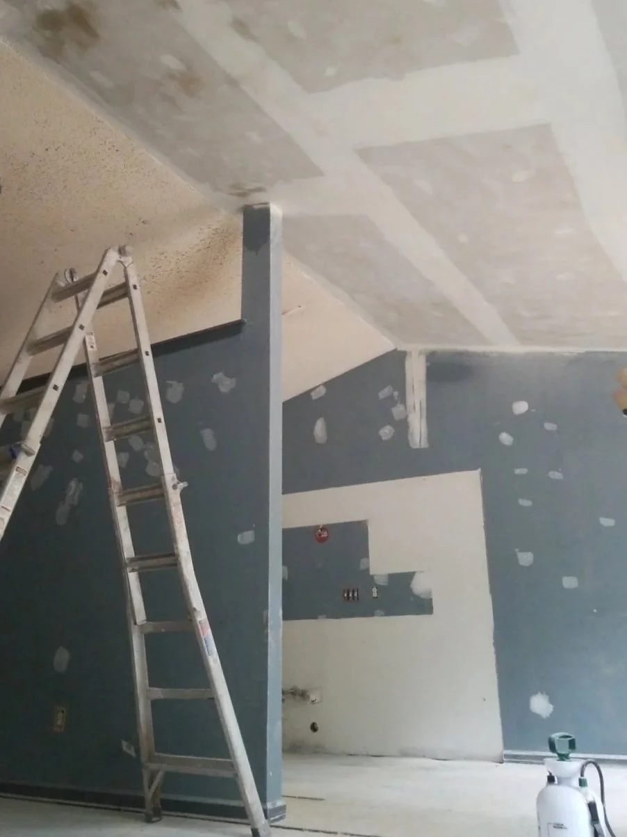 Interior of a building under renovation with drywall patches on blue and white walls and ceiling, a metal ladder leaning against a wall, and a fire extinguisher in the corner.