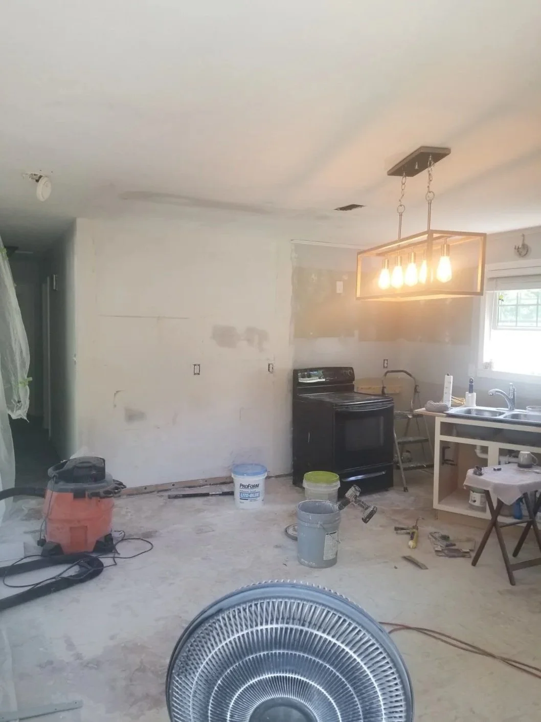 Kitchen undergoing renovation with tools, paint, and construction materials scattered around, and a modern light fixture hanging from the ceiling.