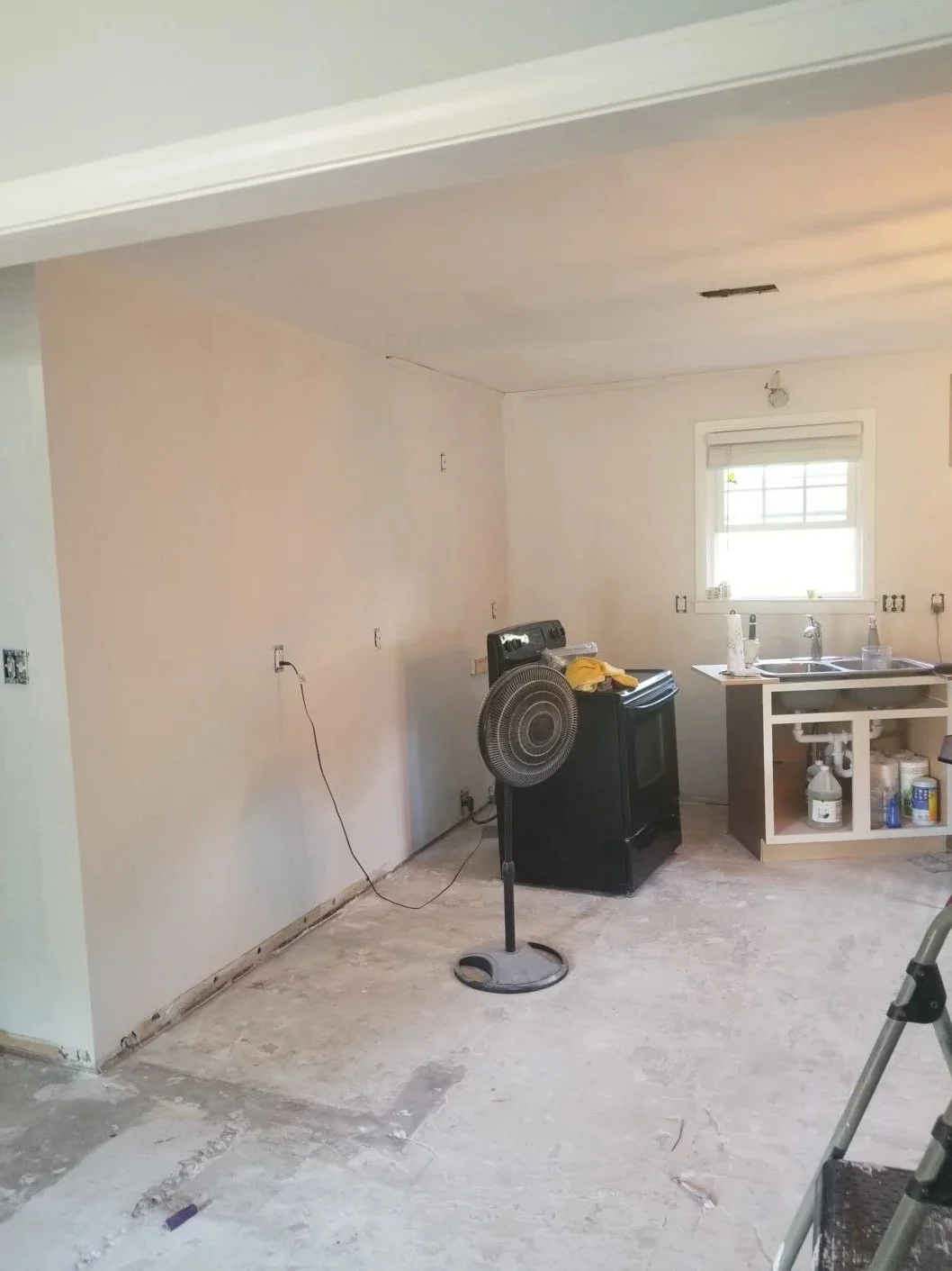 An unfinished kitchen with construction tools including a standing fan, a stove, and a sink with uninstalled cabinetry. The walls are partially painted, and the floor is bare.