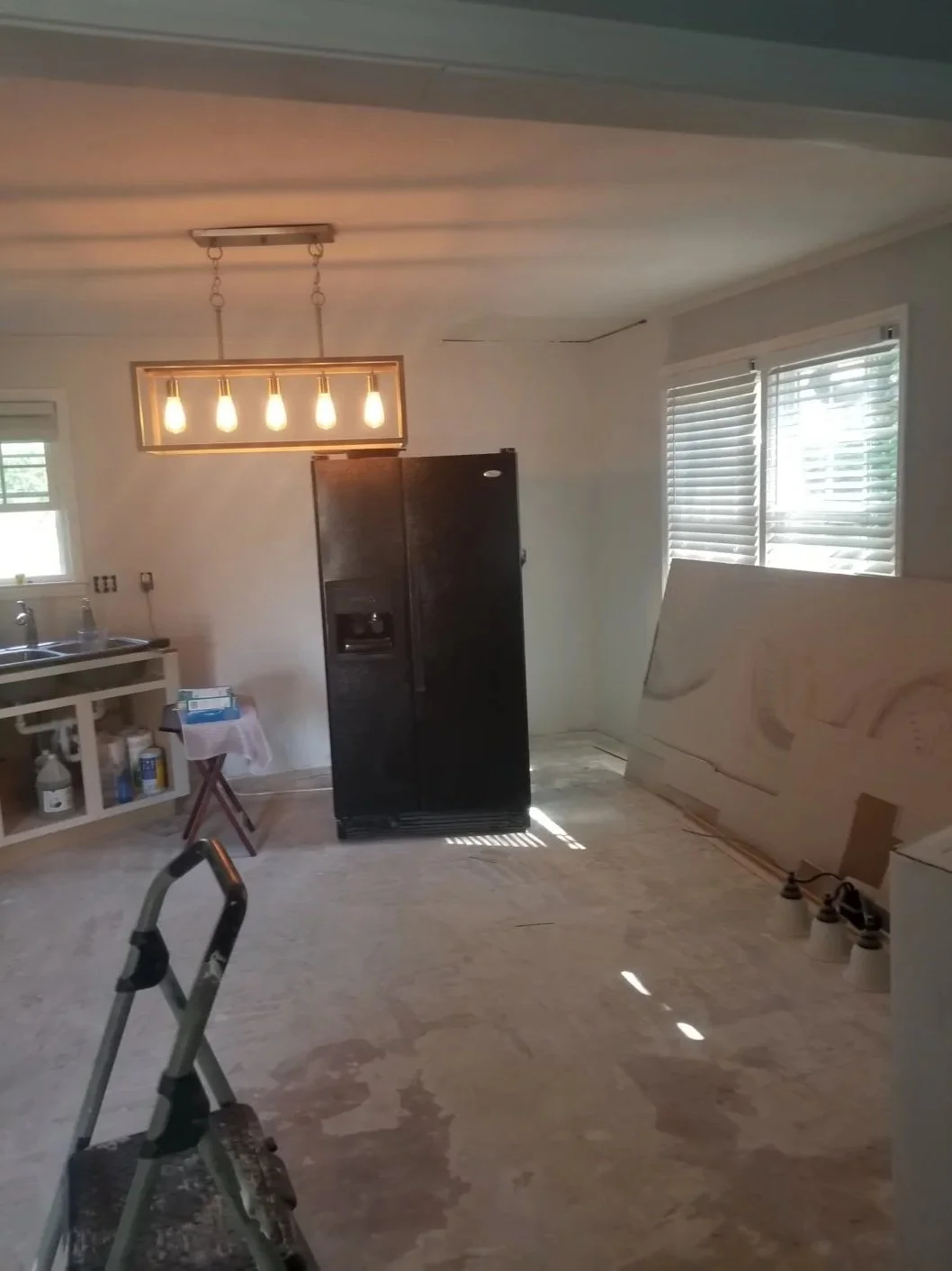 Living room under renovation featuring a ladder, black refrigerator, window blinds, and a ceiling light fixture with five bulbs.