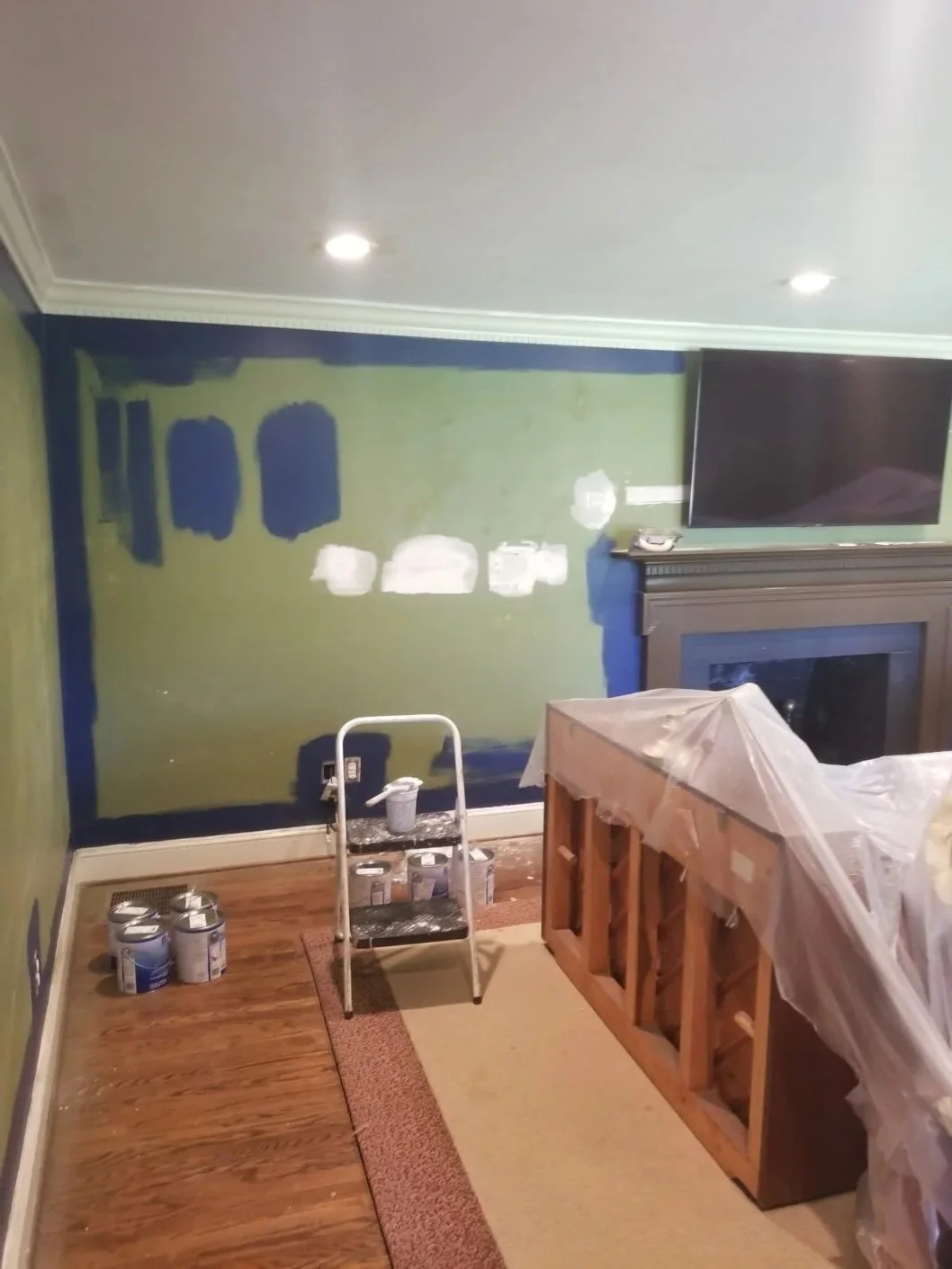 Living room undergoing wall painting with green and blue paint, paint cans on the floor, a small step ladder, a fireplace, and a mounted television.