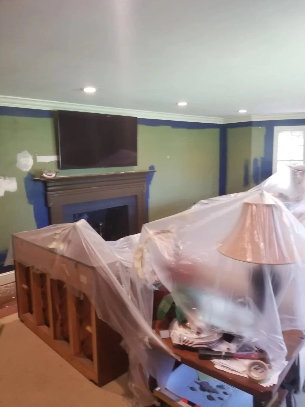 Living room undergoing renovation with green walls, a fireplace, and a mounted television. Furniture is covered with plastic sheeting.