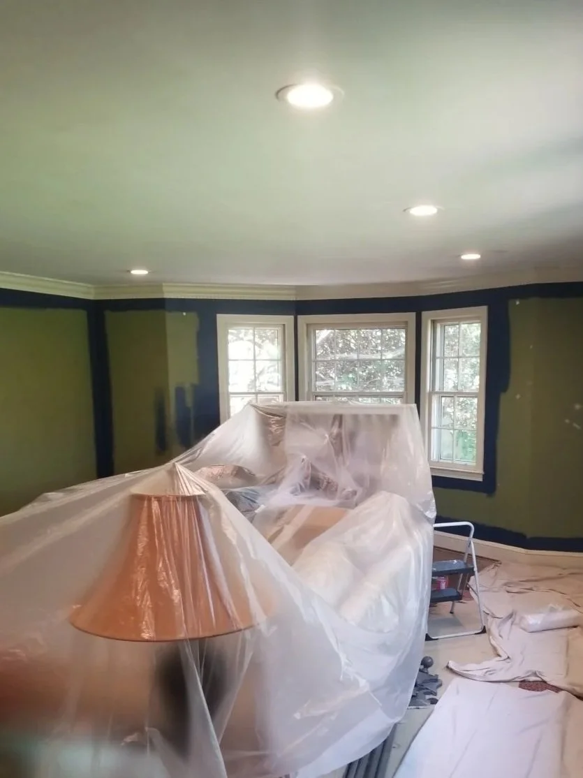 Living room undergoing renovation, covered with plastic sheets, with lamps, window, and a step ladder.