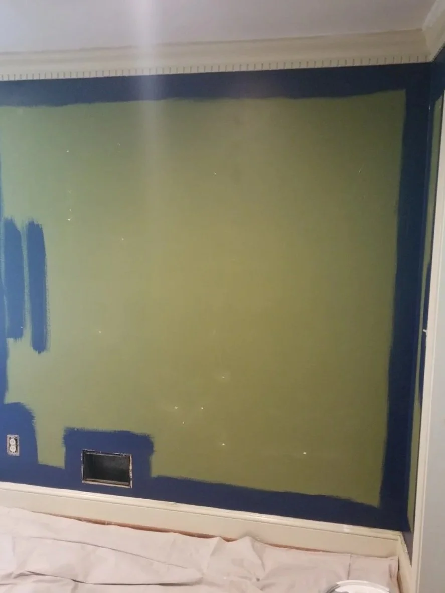 A wall with partially painted sections, with blue paint around the edges and a large central area painted in yellow-green. There are some paint gaps and a vent near the bottom of the wall. A sheet or drop cloth is partially visible on the floor.
