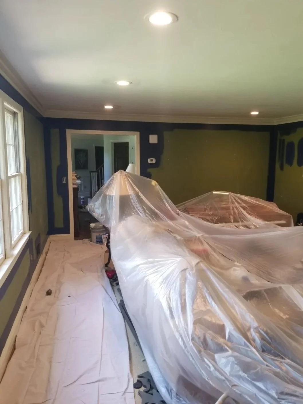 Living room undergoing renovation with plastic coverings over furniture, ceiling lights on, and a window on the left.