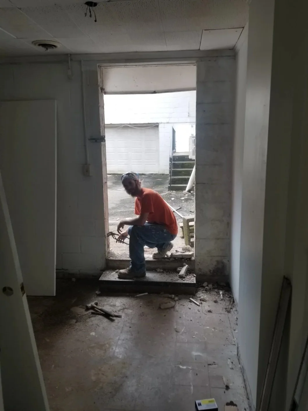 A man squatting on a small step outside a building under construction or renovation, working with tools, through a doorway leading outside to a concrete yard with stairs, cinder blocks, and construction debris.