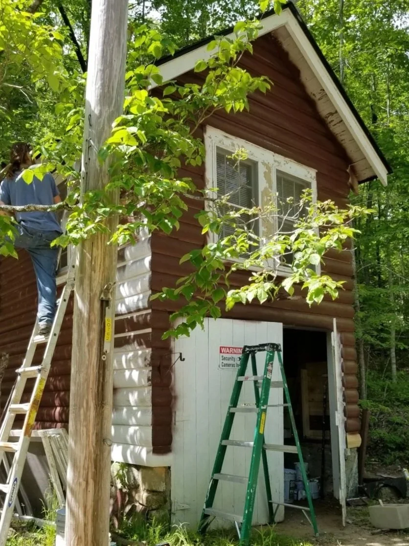 A small two-story wooden house under renovation, with a person on a ladder working on the exterior wall, surrounded by green trees and foliage.
