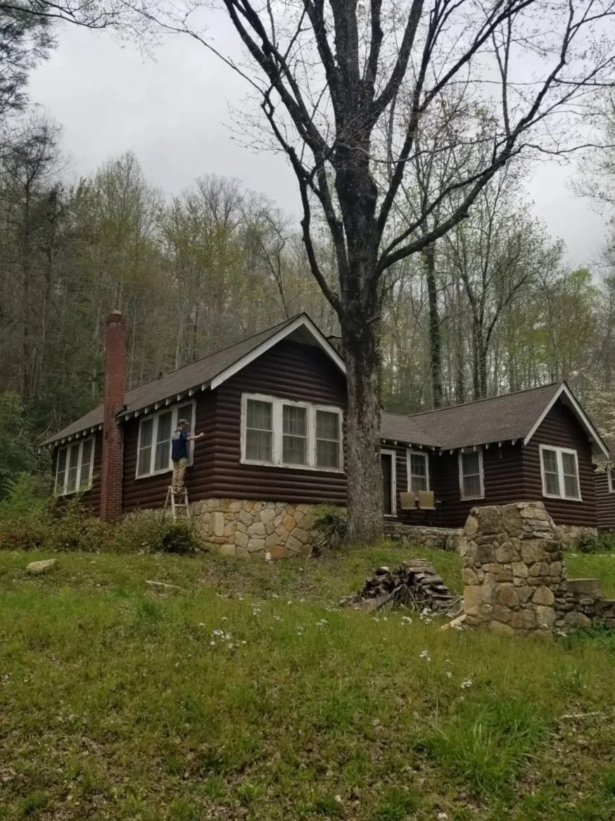 A rustic house with dark wood siding, multiple windows, and stone foundation, surrounded by trees and a grassy yard. A person stands on a ladder near a chimney.