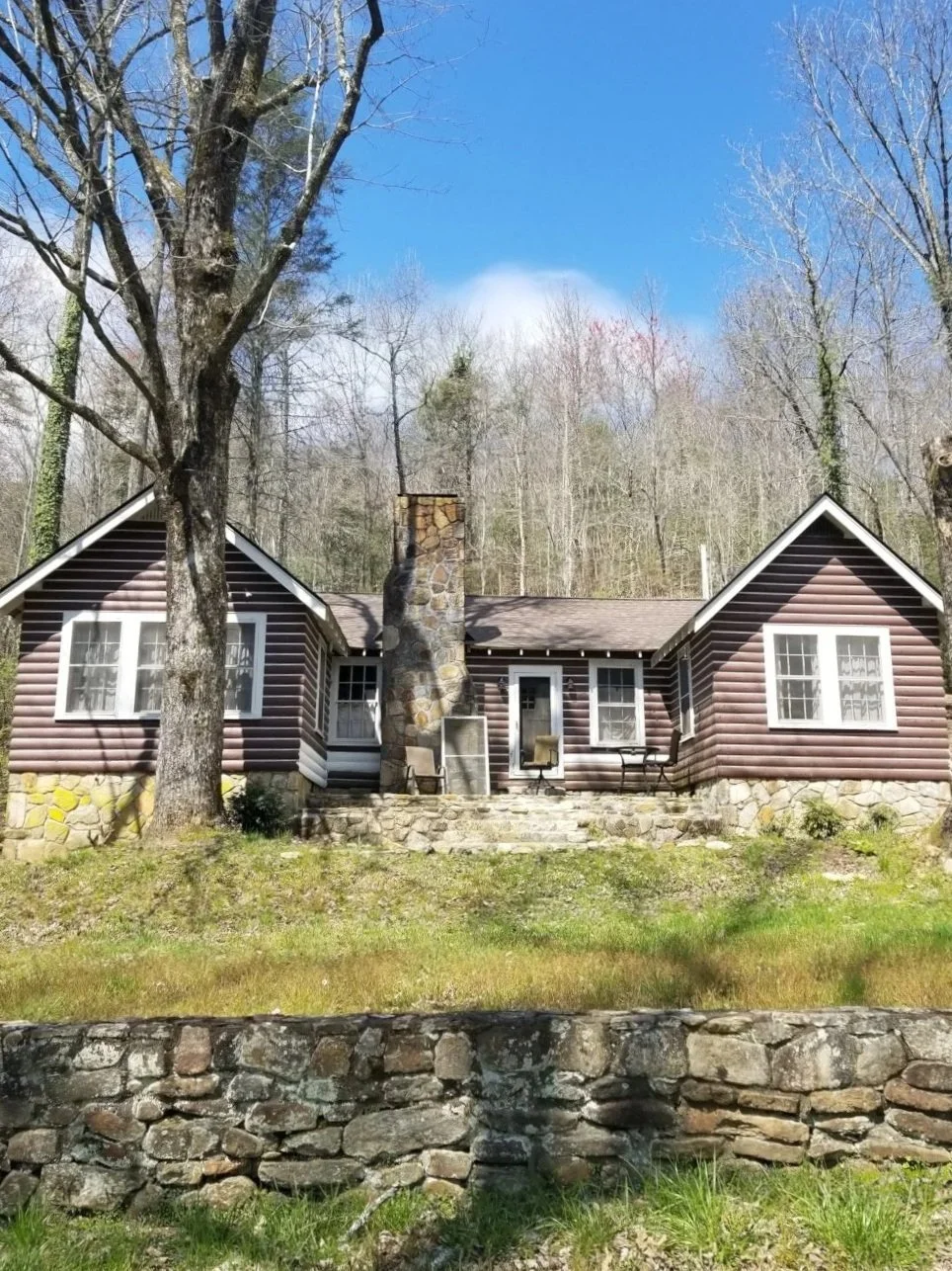 A rustic house with brown logs, white trim, and a stone chimney, surrounded by trees and a stone wall in front.