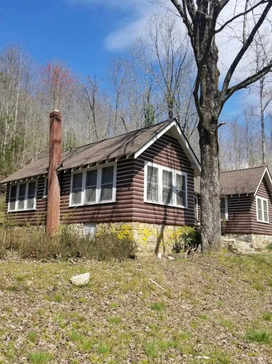 A cozy log cabin with white-framed windows and a stone foundation, situated on a wooded hillside with a large, leafless tree in front, under a partly cloudy blue sky.