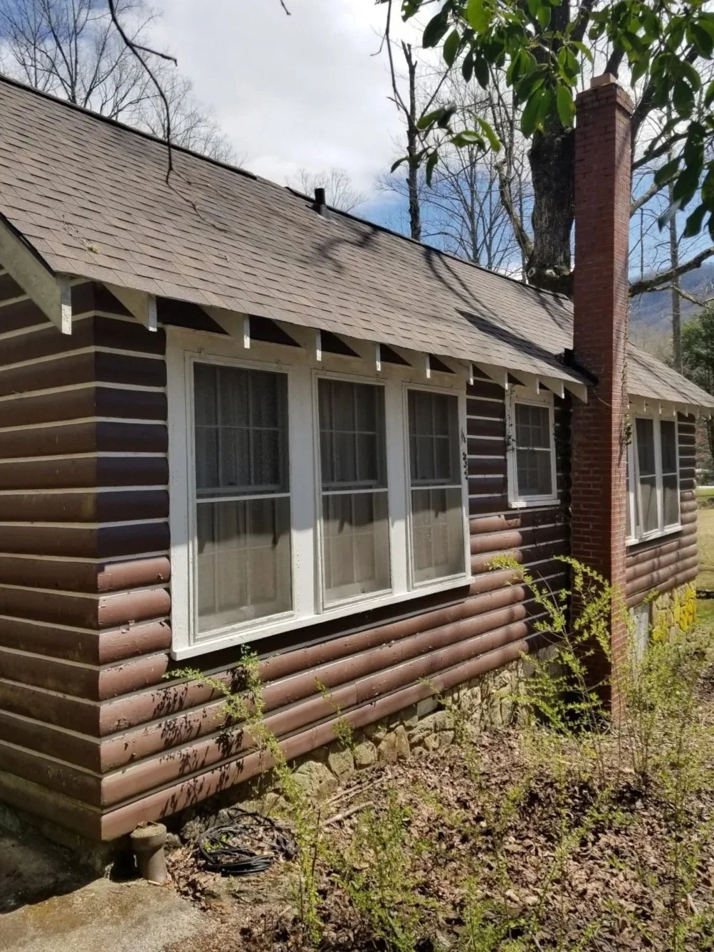 A house with brown log cabin siding, white-framed windows, a sloped shingled roof, and a large brick chimney on the side, surrounded by trees and plants.