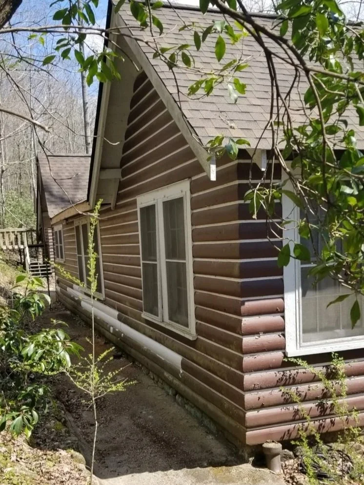 A brown log cabin with white trim on the windows, surrounded by green trees and bushes.