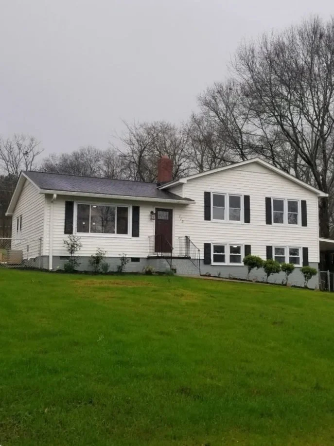 White two-story house with black shutters and a front porch, surrounded by a green lawn and leafless trees in the background on a cloudy day.