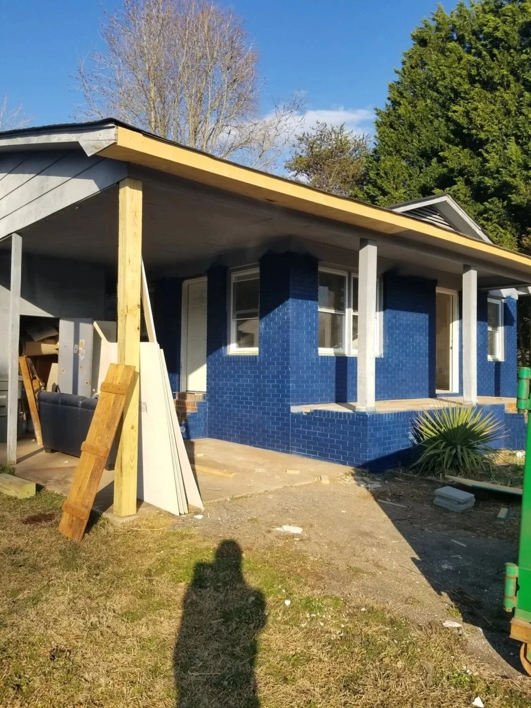 A house under construction with blue brick walls, white window frames, and a partially finished roof with wooden support beams. Construction materials and tools are visible nearby.