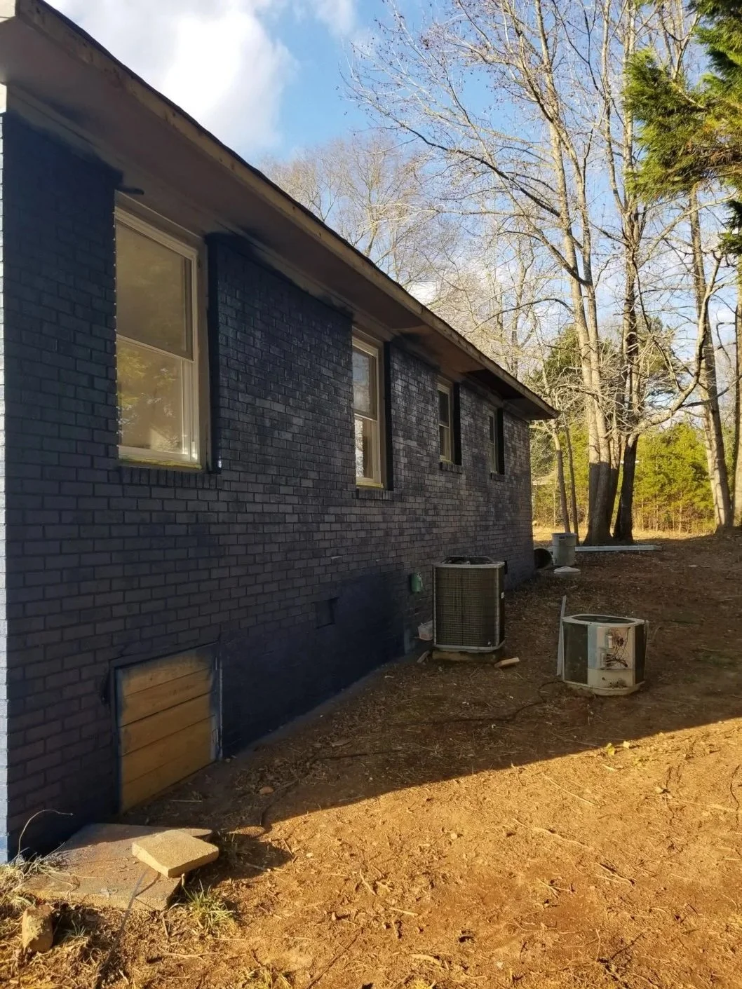 Backyard with a black brick house, several windows, and trees in the background. Outdoor HVAC units and construction debris in the dirt yard.